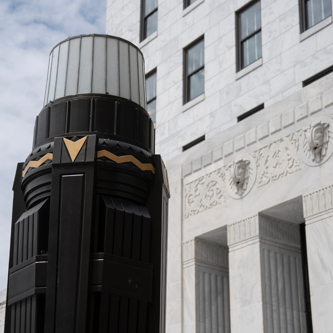 Close-up of an art deco-style black and gold street light with a frosted glass top, set against a white marble building facade featuring intricate carvings.