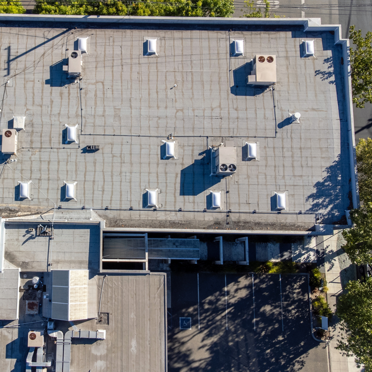 An aerial, top-down drone photograph of a flat, grey commercial rooftop with multiple rectangular skylights and several HVAC units casting long shadows in the sunlight.