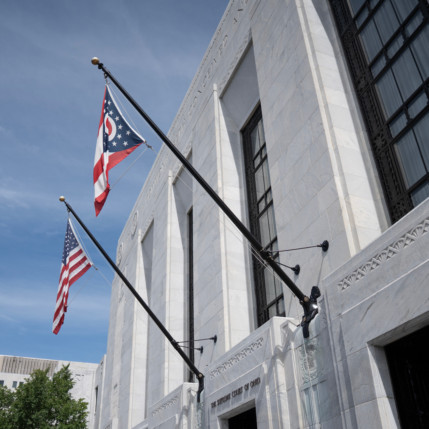 An angled, upward-facing shot of the white marble facade of The Supreme Court of Ohio building under a clear blue sky, featuring the American flag and the swallowtail Ohio state flag flying from black flagpoles mounted over the entrance.