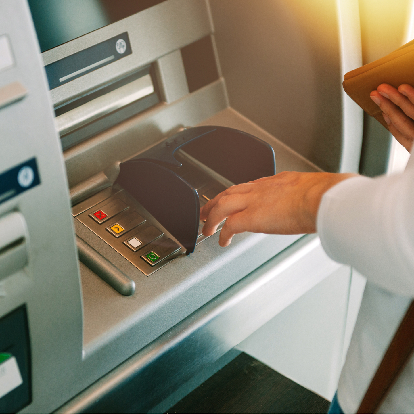 A person's hand entering a PIN on an ATM keypad covered by a dark privacy shield.