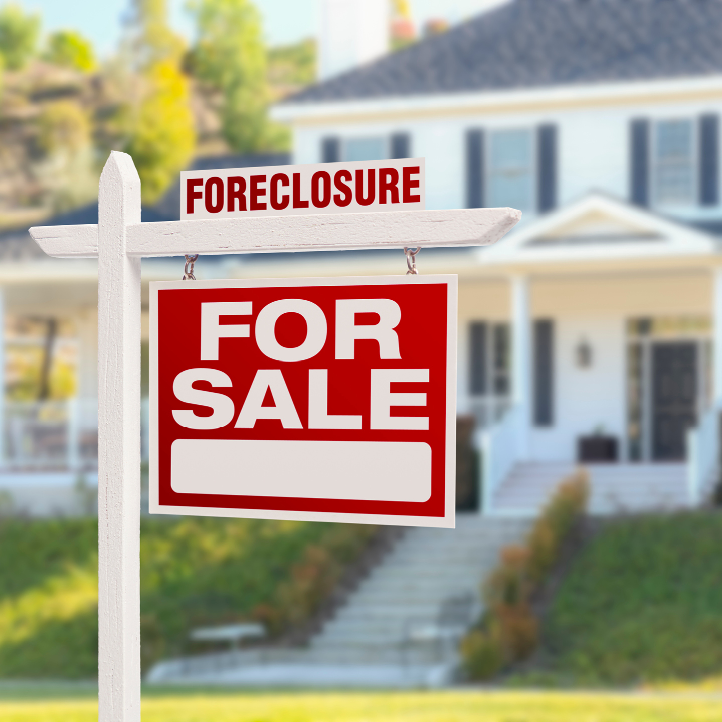A white real estate signpost stands in front of a blurry, two-story white house with a green lawn. The main sign is red with bold white text that reads 'FOR SALE' and includes a blank white rectangle at the bottom for contact details. A smaller white rider sign sits on top of the post with the word 'FORECLOSURE' printed in bold red capital letters.