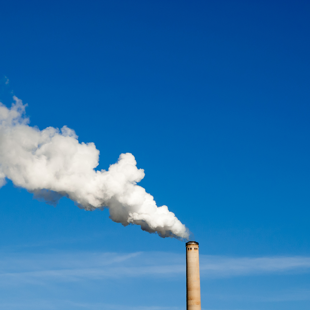 A tall industrial smokestack releasing a thick plume of white smoke into a clear, bright blue sky