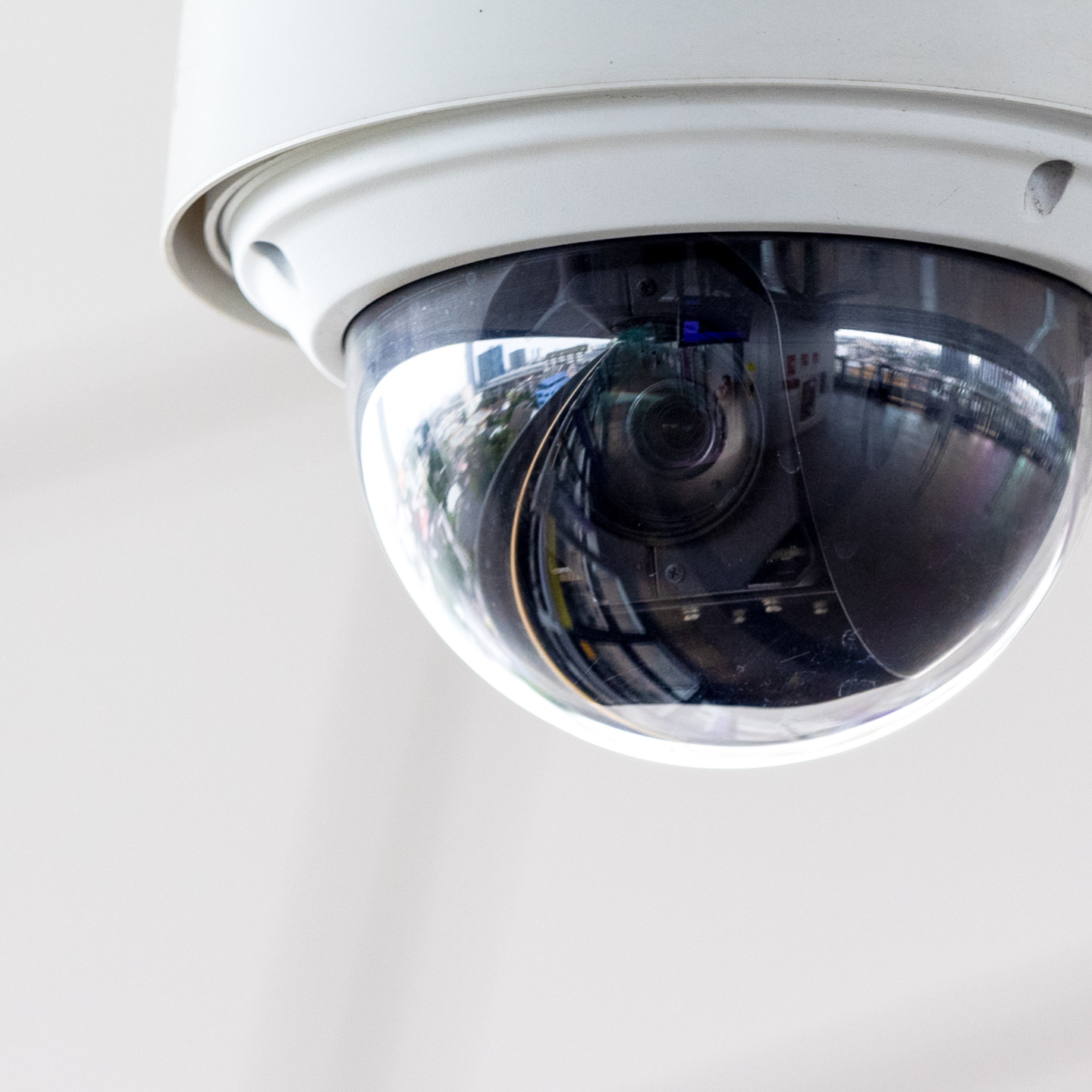 An outdoor dome security camera is mounted to a white structure, featuring a clear glass housing that captures a distorted reflection of its urban surroundings.
