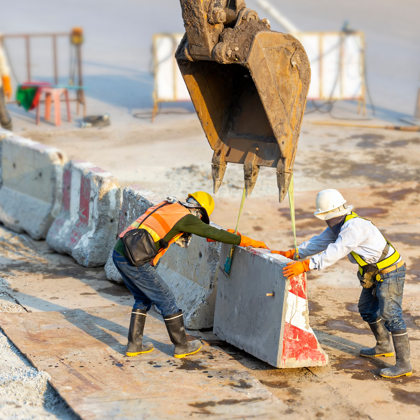 An orange-vested worker wearing blue jeans and black boots guides a large concrete barrier being lifted by an excavator bucket, while a white-helmeted worker balances the other side.