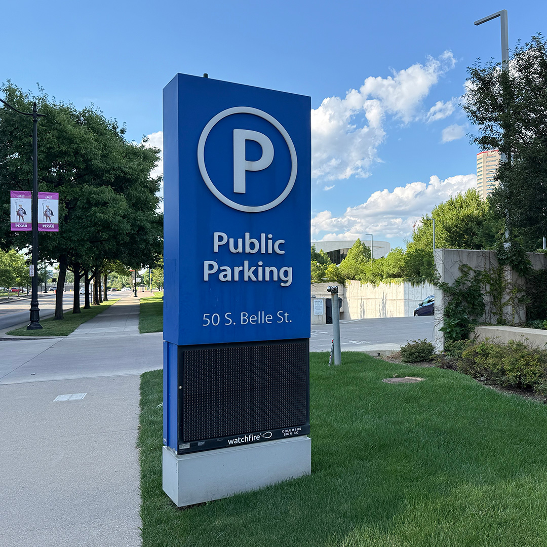 A large blue public parking sign displaying a white letter 'P' in a circle, reading 'Public Parking' and '50 S. Belle St.' above a black electronic display screen, set outdoors next to a sidewalk and green grass on a sunny day