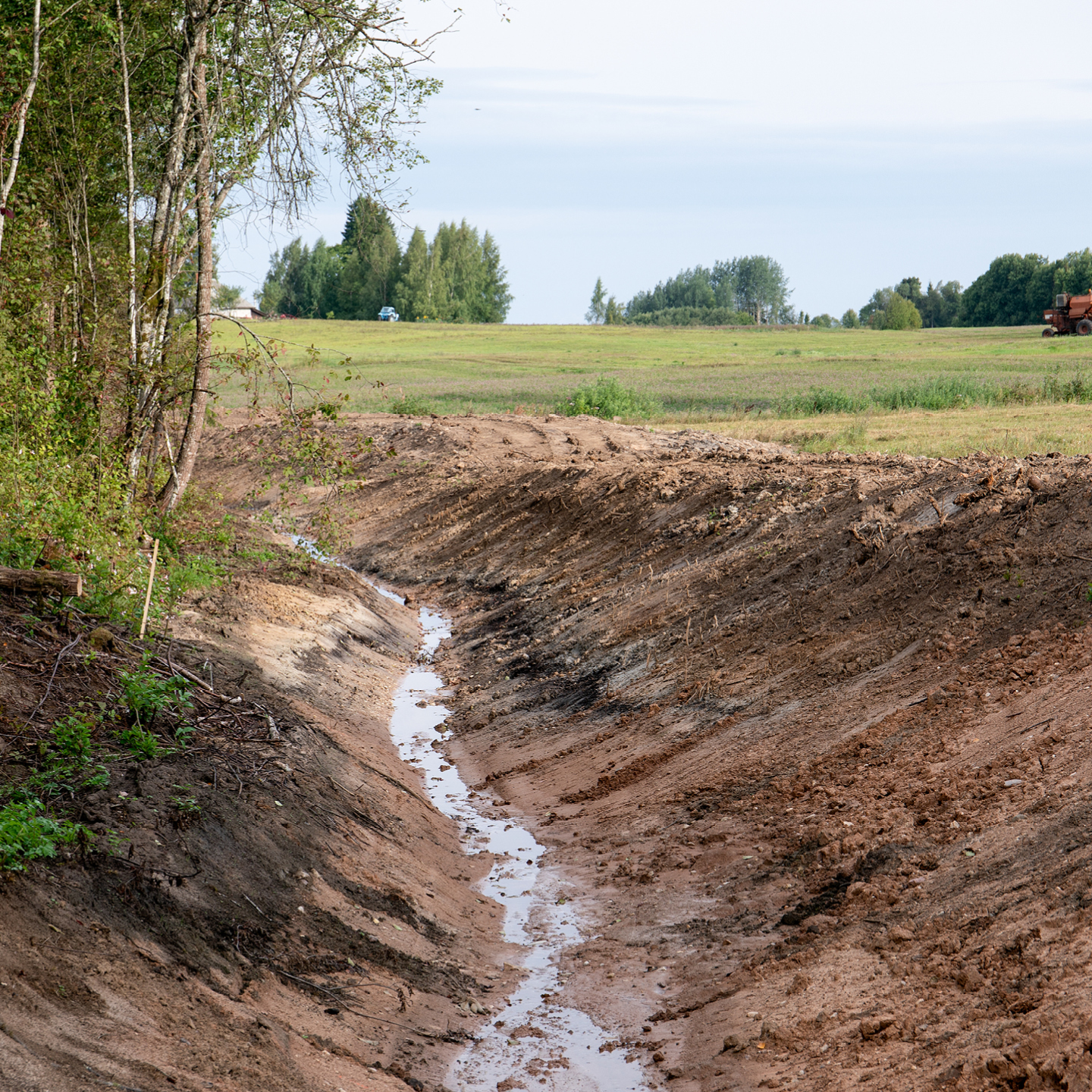 A newly excavated drainage ditch running down a gentle slope in a rural field. A small stream of water flows along the muddy bottom of the ditch, which features sloped, dirt banks. A grassy field and a line of distant trees are visible under a pale sky.