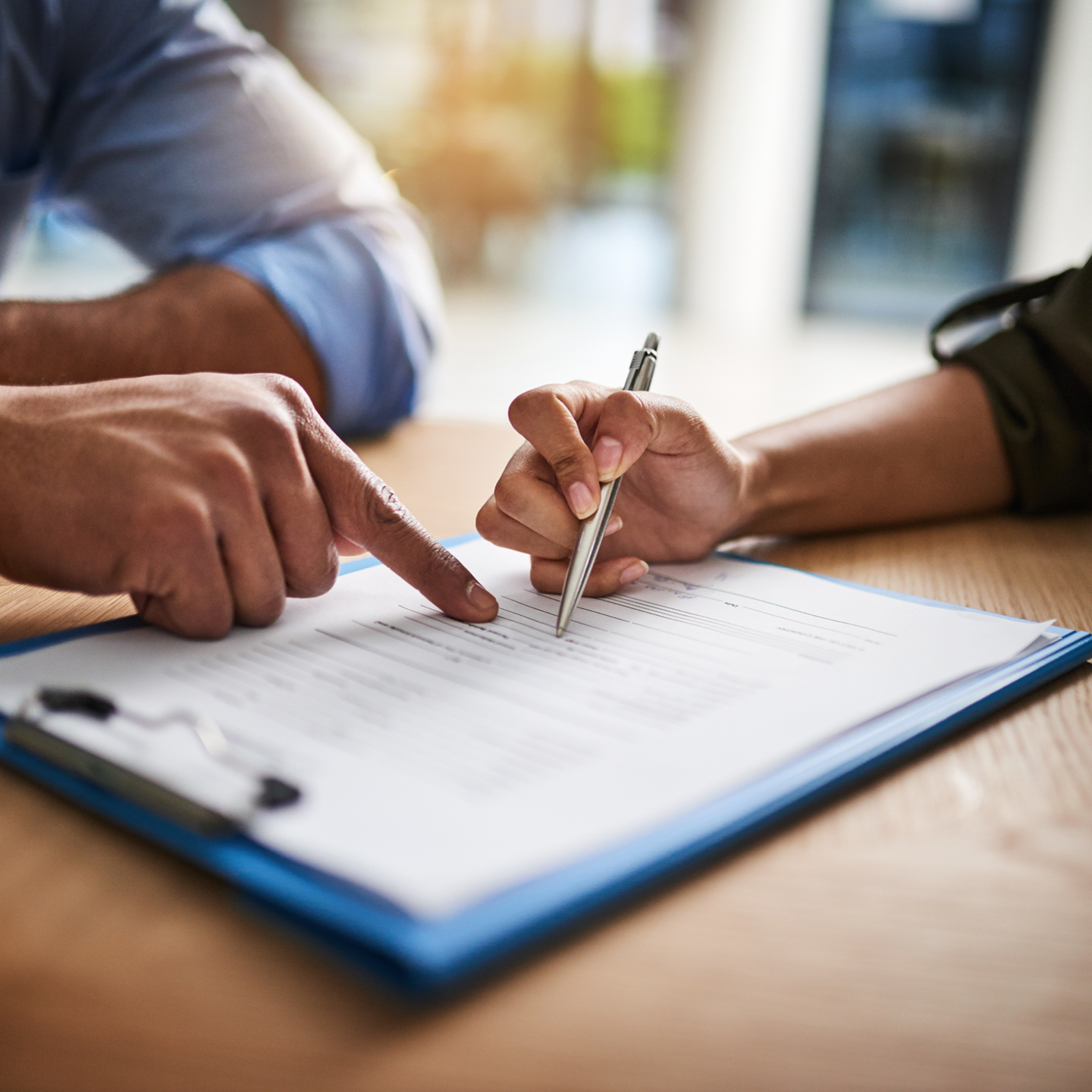 A hand points to a section on a clipboard document while another hand uses a silver pen to sign it.