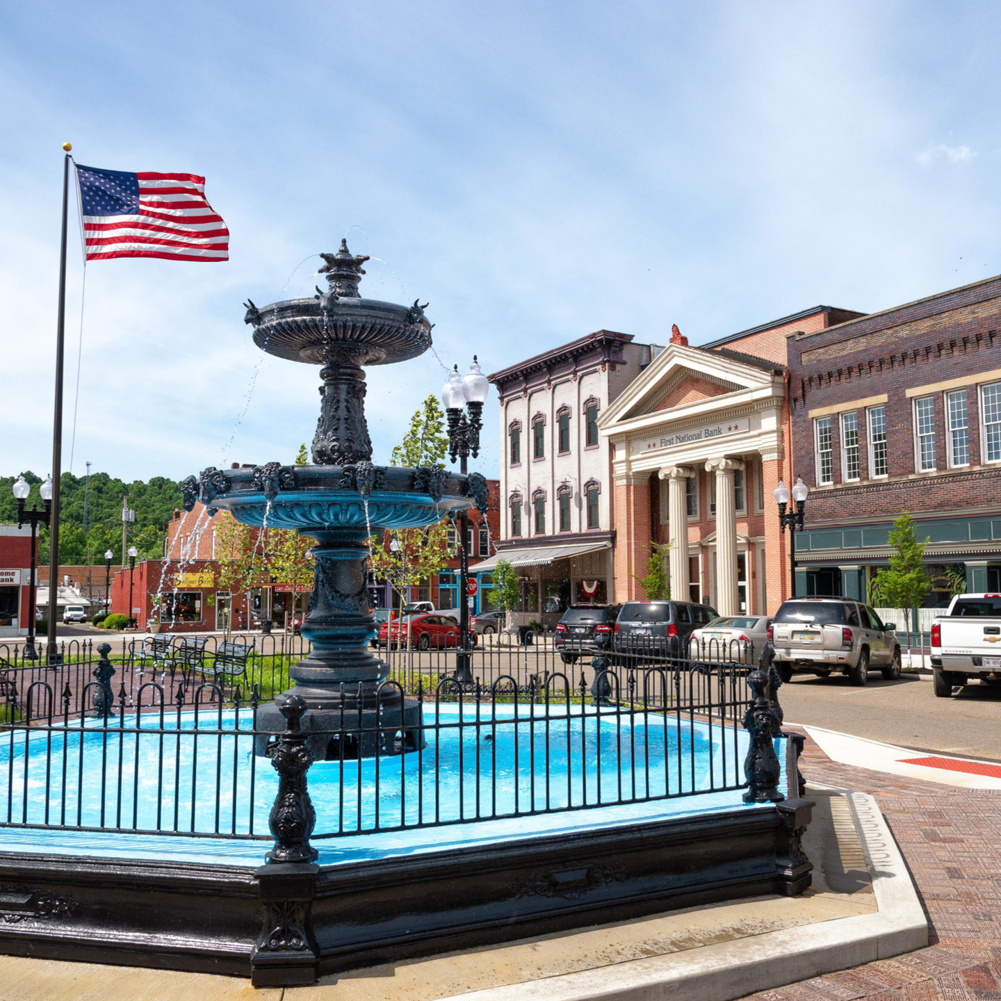 A historic town square featuring a large, ornate black tiered fountain with bright blue water inside a black metal fence.