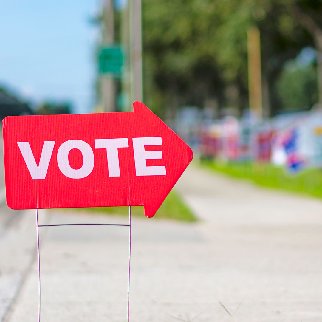 A large, red, arrow-shaped yard sign with the word 'VOTE' printed in bold, white capital letters stands on thin metal wire legs along a concrete sidewalk, pointing toward the right.