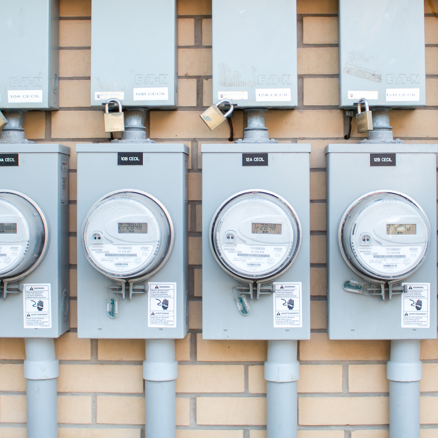 Four gray electrical meters mounted on a brick wall, with digital displays and labels indicating identifiers, some secured with padlocks.