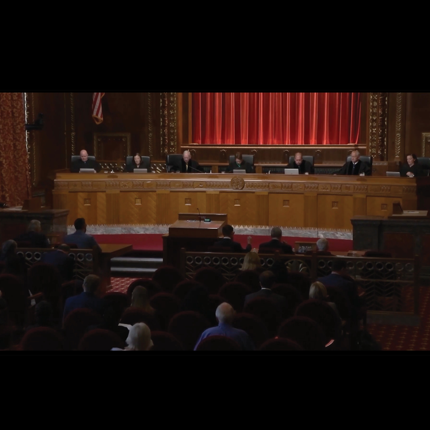 A court room with red chairs and a large wooden bench with seven justices.