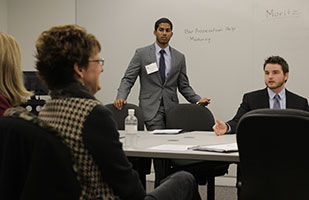 Image of two men in suits, one standing, one seated at a table, addressing a group of people