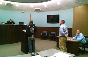 Image of a man standing at a podium in a courtroom addressing a judge; another man stands behind him next to a table where a third man sits