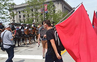 Image of a man with a flower tucked behind his ear holding a red flag in front of a line of mounted police