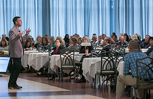 Image of a man standing in front of a roomful of people sitting at tables giving a presentation