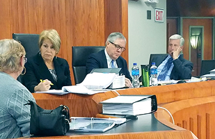 Image of a woman and two men seated on a courtroom bench listening to another woman speak