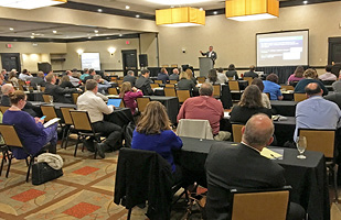 Image of a roomful of people listening to a speaker standing at a podium