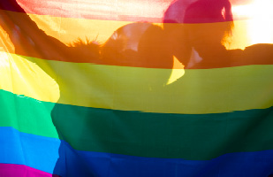 Image of the rainbow LGBT pride flag in front of a silhouette of two people (DusanManic/iStock)