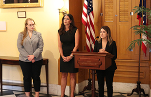 Image of a woman speaking from a podium with two additional women standing to her left