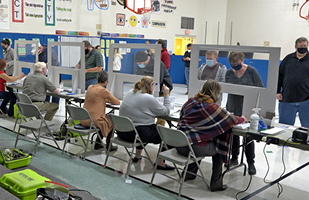 Image of people sitting at tables behine plexiglass barriers assisting voters