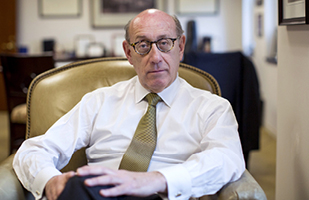 Image of a man wearing glasses, a dress shirt, and tie, sitting in a chair