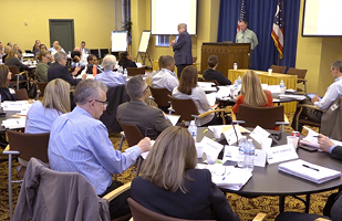 Image of men and women sitting at tables listening to a man speaking from a podium