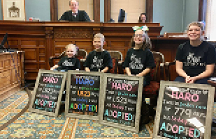 Image of three girls and a boy sitting in chairs in a courtroom smiling and holding signs declaring they have been adopted, while a male judge smiles down from the bench