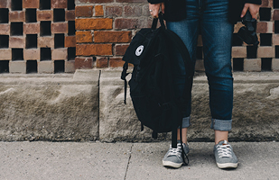 Image of the lower half of a person in jean and gray athletic shoes, holding a black backpack and standing on a cement sidewalk