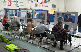Image of an election polling site, with five people sitting at tables and several people standing in front of the tables