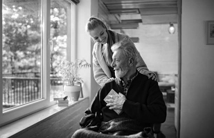 Image is of an older man in a wheelchair with a younger woman behind the wheelchair, leaning over to talk with him (iStock/VorDa)