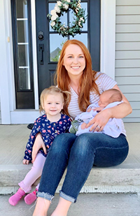 Image of a smiling woman sitting on front porch steps holding an infant in one arm and her other arm around a toddler