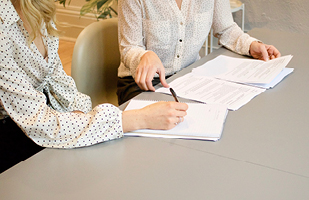 Image of two women sitting next to each other at a table with paperwork in front of them