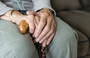 Image of the legs and hands of a person sitting in a chair appearing to be an elderly woman who is holding a cane in her lap