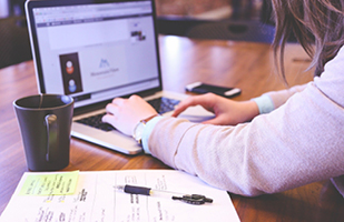 Image shows the arms and hands of a person working on a laptop computer with a coffee cup and papers in the foreground