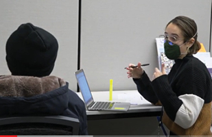 Image show a woman sitting at a table in front of a laptop turning to her left to talk to a person on her left whose back is seen with a winter coat and hat