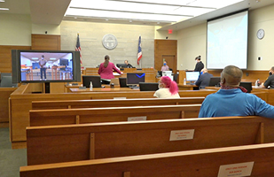 Image of a courtroom from the back looking forward with the backs of two people sitting in the audience, one person standing facing the judge who is on the bench with a large video screen next to the person who is standing