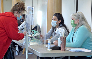 Image of two women sitting at a table behind a plexiglass window assisting another woman with voting