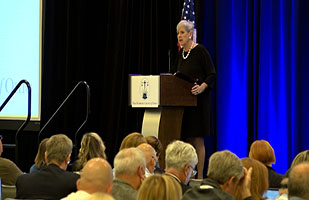 White woman with gray hair on a podium behind a lectern speaking to hundreds of people in a room.