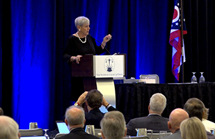 A white woman with gray hair on a stage behind a lectern and microphone speaks to hundreds of people in a room.