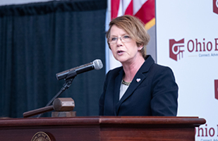 Woman wearing a blue suit speaking from a wooden podium to a room full of men and women.