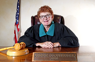 Image of a female judge with red hair and wearing a black judicial robe seated at a wooden table with a wooden gavel at her side.
