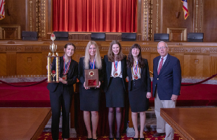 Image of five girls, all wearing blue suits and medals around their neck, standing next to a man, also wearing a blue suit, in the courtroom of the Thomas J. Moyer Ohio Judicial Center. The first girl on the left is holding a large trophy and the second girl on the left is holding a plaque.