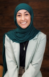 Image of a smiling woman wearing a green hijab, black top, and beige blazer.