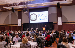 Image of a large room full of men and women sitting at round tables while a person speaks from behind a podium.