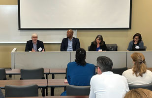 Image of two men and two women sitting side-by-side at a long, enclosed table speaking to an audience of men and women.