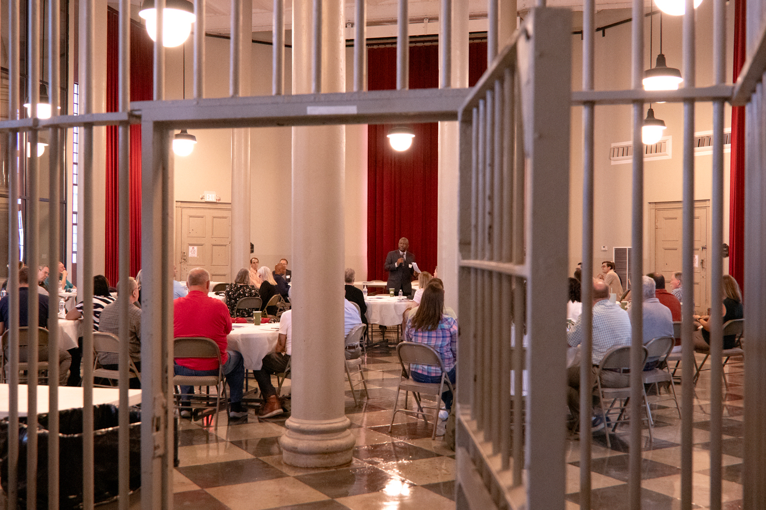 A man wearing a black suit and holding a microphone speaks to a group of men and women seated at round tables. The image is seen through an open jail cell door.