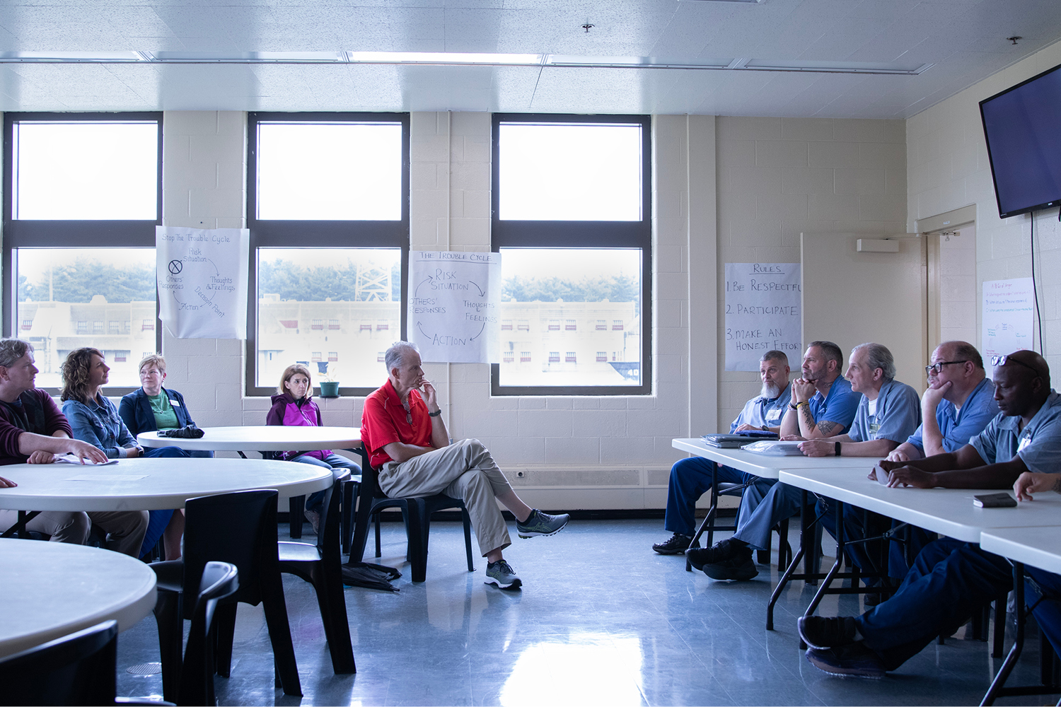 A man wearing a red, short-sleeve shirt sits with his legs crossed at a table with four women all listening to a group of men wearing blue prison clothes seated at long, white folding tables.