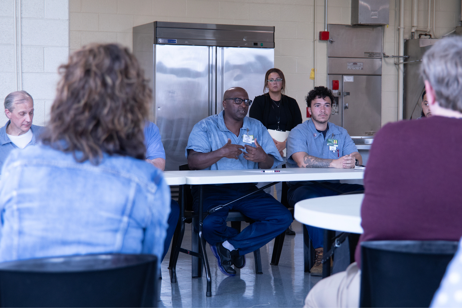 A group of men and women seated at round tables listen to a man wearing jeans and a blue prison shirt speak.