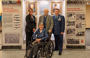 A woman wearing a blue suit stands next to a man wearing a grey blazer and another man wearing an Air Force dress uniform. All are standing behind a woman seated in a wheelchair.
