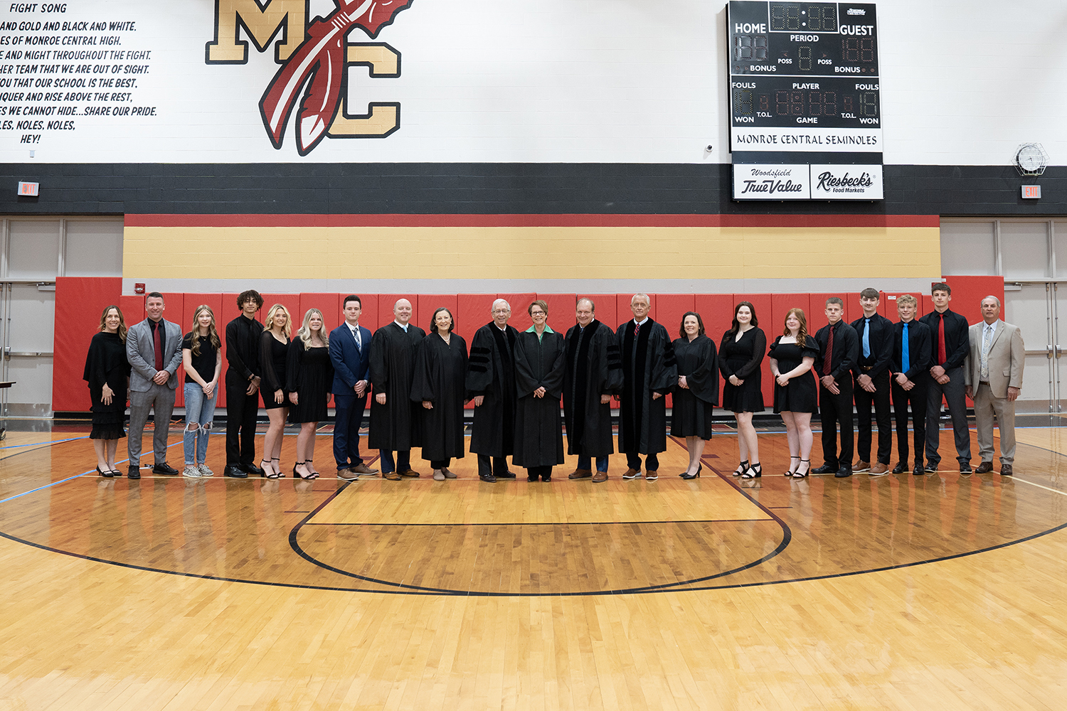 Seven male and female judges wearing black judicial robes stand at the center of a single line of teachers and students in a gymnasium.